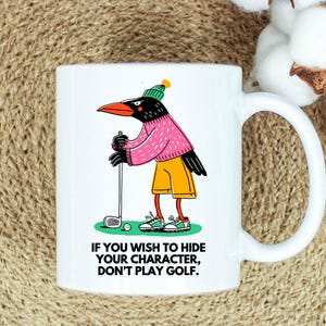 May include: White ceramic mug featuring a cartoon crow in a pink sweater, yellow shorts, and a green beanie, playing golf. The mug has the text: "IF YOU WISH TO HIDE YOUR CHARACTER, DON'T PLAY GOLF."