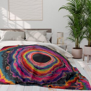 Puede incluir: Una manta con un patr&oacute;n abstracto circular y multicolor vibrante con anillos conc&eacute;ntricos de color rojo, naranja, amarillo, verde, azul y morado que irradian desde un centro negro s&oacute;lido, colocada sobre una cama con ropa de cama blanca.
