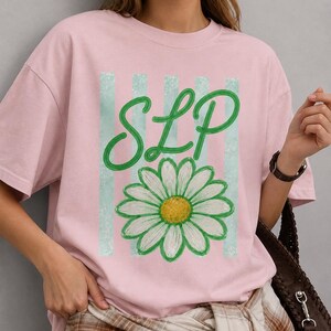 Puede incluir: Una camiseta de manga corta de color rosa claro con un estampado gráfico de las letras SLP en cursiva verde sobre una margarita blanca con centro amarillo, sobre un fondo de rayas verticales de color azul claro en la parte frontal.