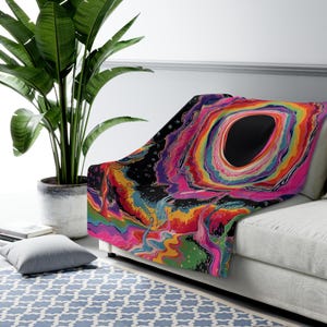 Puede incluir: Una manta suave con un dise&ntilde;o abstracto psicod&eacute;lico y vibrante, con patrones de remolinos en colores rosa ne&oacute;n, naranja, amarillo, azul y morado que rodean una gran forma ovalada de color negro s&oacute;lido sobre un fondo oscuro.