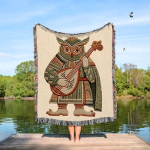 May include: A woven tapestry blanket featuring an owl playing a lute. The owl is depicted in shades of green, brown, and beige, with intricate details. The blanket has a cream-colored background and a fringed edge. The scene is set outdoors near a lake.