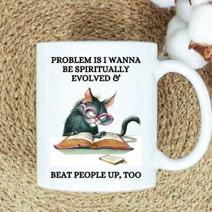 May include: White ceramic mug featuring a cartoon cat wearing glasses and reading a book. The mug has the text "PROBLEM IS I WANNA BE SPIRITUALLY EVOLVED & BEAT PEOPLE UP, TOO."