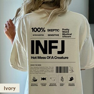May include: A beige t-shirt with a black graphic design that says "100% Skeptic", "Introverted", "Sensitive", "Pretty Weird Mystical Unicorn", "INFJ", "Hot Mess Of A Creature", and includes icons for "Socially Selective", "Fantasy Lover", "Intuitive Insightful", "Easily Hurt", and "Walking Paradox".