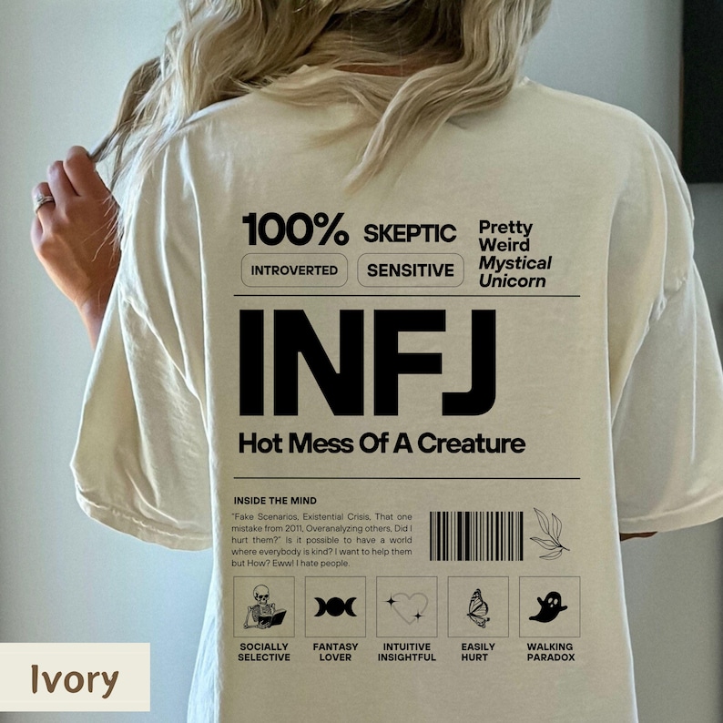 May include: A beige t-shirt with a black graphic design that says "100% Skeptic", "Introverted", "Sensitive", "Pretty Weird Mystical Unicorn", "INFJ", "Hot Mess Of A Creature", and includes icons for "Socially Selective", "Fantasy Lover", "Intuitive Insightful", "Easily Hurt", and "Walking Paradox".