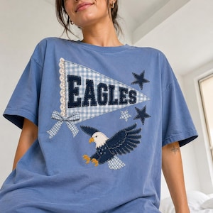 May include: A blue short-sleeve t-shirt featuring a central pennant graphic with the text EAGLES in dark blue letters. The design includes a white and blue gingham pattern, a small gingham bow, a flying eagle, and three dark blue stars.
