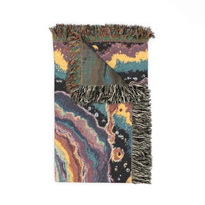Puede incluir: Una manta tejida con un patr&oacute;n abstracto multicolor con capas arremolinadas de p&uacute;rpura, verde azulado, amarillo y naranja. La manta tiene una esquina doblada que revela el reverso y est&aacute; terminada con flecos decorativos en los bordes.