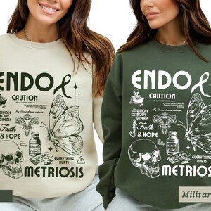 May include: Two sweatshirts with a graphic design featuring a butterfly, skull, and uterus. The text on the sweatshirts reads "Endo Caution" and "Metriosis". The sweatshirts are available in sand and military green.