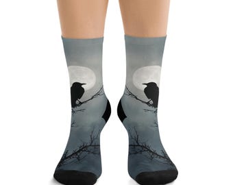 May include: A pair of crew socks featuring a spooky design. The socks display a silhouette of a raven perched on a branch in front of a full moon. The background is a gradient of gray and blue, creating a night sky effect.