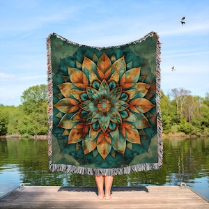 May include: A woven throw blanket featuring a large, stylized floral design in shades of teal, orange, and gold. The blanket has a dark green background and fringed edges. The person holding the blanket is standing on a wooden dock.