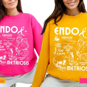 May include: Two pink and gold sweatshirts with a white graphic design. The design features a butterfly, a skull, and the text "ENDO CAUTION EVERYTHING HURTS METRIOSIS".
