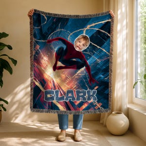 May include: A woven throw blanket featuring a Spiderman-themed design with a child's face superimposed on the superhero. The blanket has a cityscape background and the name "CLARK" in large, blue letters. The blanket has a fringed edge.