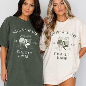 Puede incluir: Dos camisetas holgadas, una verde oliva y otra crema, con el texto "BITCHES & BEACHES" sobre un gráfico de dos figuras y el texto "FERAL CLUB HAWAII". Las camisetas tienen un aspecto vintage y desgastado.