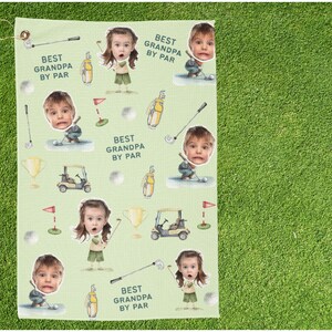 May include: A light green golf towel featuring a repeating pattern of a child's face, golf equipment, and the text "BEST GRANDPA BY PAR". The design includes golf clubs, golf balls, golf carts, and trophies.