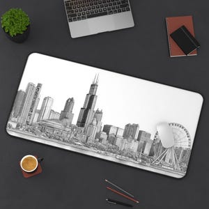 May include: A white rectangular desk mat featuring a detailed black and white illustration of the Chicago skyline, including the Willis Tower and a Ferris wheel. A computer mouse rests on the mat. Other items include a laptop, notebook, and a cup of coffee.