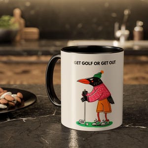 May include: A white ceramic mug with a black interior and handle. The mug features a cartoon crow dressed in golf attire, holding a golf club. The text "GET GOLF OR GET OUT" is printed on the mug.