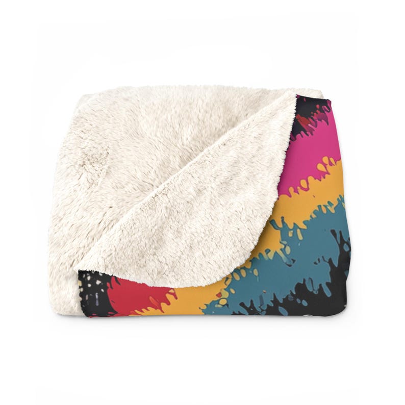 Puede incluir: Una manta doblada que presenta un forro de vell&oacute;n sherpa texturizado de color crema en un lado y un patr&oacute;n abstracto de salpicaduras de pintura multicolor en el otro, mostrando rayas diagonales de color rosa intenso, amarillo, azul verdoso y negro.