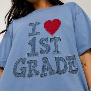 May include: A light blue t-shirt featuring a red fabric heart and the text "I 1ST GRADE" appliqued in a frayed denim fabric. The letters and numbers have a textured, distressed appearance with visible stitching around the edges of each character.