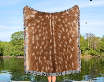 May include: A brown and white throw blanket with a deer-like pattern is held up against a blue sky and lake background. The blanket has a fringed edge with multiple colours. The blanket is made of a soft material.