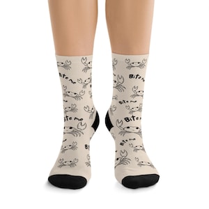 May include: A pair of beige socks with a repeating pattern of cartoon crabs and the text "Bite me" in black. The socks have black heels and toes.