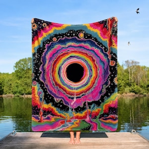Puede incluir: Una manta rectangular con un dise&ntilde;o psicod&eacute;lico vibrante que presenta un gran c&iacute;rculo negro central rodeado de anillos conc&eacute;ntricos de colores brillantes rosa, naranja, amarillo, verde y azul, sobre un fondo de espacio oscuro y estrellado.