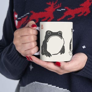May include: A white ceramic mug featuring a black illustration of a frog and a cat. The frog has a large open mouth, and the cat is holding a small bowl. The mug is held by a person wearing a navy blue sweater.
