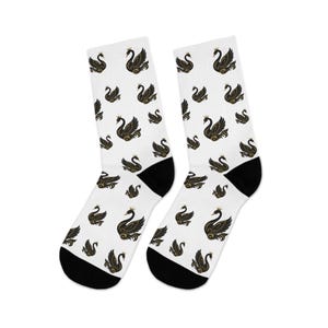 May include: A pair of white socks with a black and gold swan pattern. The socks have black heels and toes. The swans are depicted with crowns and detailed wing designs, creating a regal aesthetic.