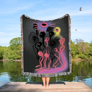 May include: A black woven throw blanket featuring a colorful, abstract illustration of figures with black silhouettes and pink and yellow accents. The blanket has a fringed edge and is held up against a lake backdrop.