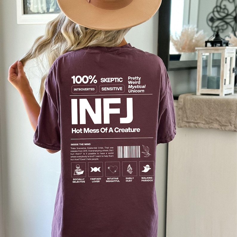 May include: A maroon t-shirt with white text that reads "100% Skeptic Introverted Sensitive Pretty Weird Mystical Unicorn INFJ Hot Mess Of A Creature". The shirt also has a graphic with a barcode and a list of personality traits, including "Socially Selective", "Fantasy Lover", "Intuitive Insightful", "Easily Hurt", and "Walking Paradox".