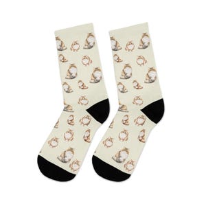May include: A pair of cream-colored socks with a repeating pattern of watercolor-style cat illustrations in shades of brown, tan, and white. The socks have black heels and toes.