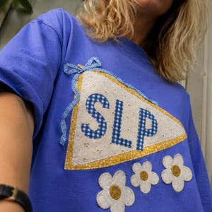 May include: A periwinkle blue t-shirt featuring a large triangular pennant appliqué with the letters SLP in blue gingham. The pennant has a yellow floral border and a blue gingham bow. Below are three white flower appliqués with yellow centers.