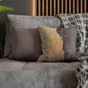 Puede incluir: Dos cojines decorativos sobre un sofá gris. Uno es liso malva, el otro tiene un diseño texturizado en tonos beige, marrón y dorado. Una manta a cuadros está sobre el sofá.
