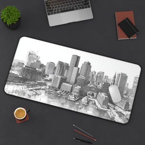 Può includere: Un tappetino per mouse in bianco e nero con un disegno dettagliato di un paesaggio urbano, probabilmente Boston, con edifici alti, acqua e barche. Un mouse per computer bianco è appoggiato sul tappetino. Altri elementi includono un laptop, un taccuino, una penna e una tazza di caffè.
