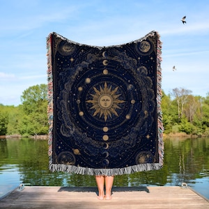 May include: A navy blue throw blanket featuring a celestial design with a sun, planets, moons, and stars. The blanket has a fringed edge with red, white, and tan accents. The design is a vintage style illustration.