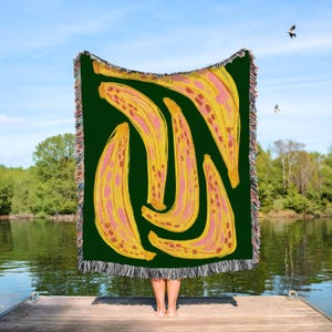 May include: A woven throw blanket featuring a dark green background with a repeating pattern of stylized yellow bananas with pink and brown accents. The blanket has a decorative fringed edge along the top and bottom, showcasing multicolored threads.