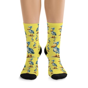 May include: A pair of yellow socks featuring a repeating pattern of a blue Shoebill bird illustration. The socks have black heels and toes. The bird has a large beak and is standing on brown boots.