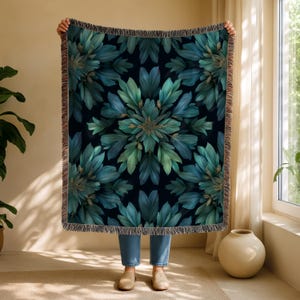 May include: A large, rectangular throw blanket with a dark navy background and a symmetrical floral design in shades of teal and green. The blanket has a fringed edge and is held up by a person.