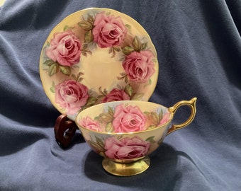 Può includere: Un set da tè vintage composto da una tazza e un piattino con un delicato motivo floreale. Il set presenta rose rosa con foglie verdi su uno sfondo crema con accenti dorati.