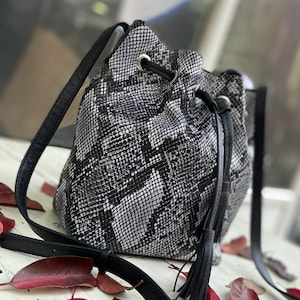Könnte beinhalten: Ein schwarz-weißes Eimer-Tasche mit Schlangenmuster, Kordelzug und schwarzen Lederriemen. Die Tasche hat einen langen Schultergurt und einen kürzeren Griff. Die Tasche steht auf einer weißen Oberfläche mit roten Blättern.