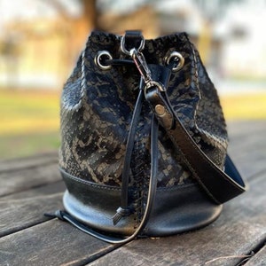 May include: A black bucket bag with a snakeskin pattern and a black leather strap. The bag has a drawstring closure and a black leather bottom.
