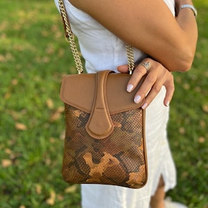 Könnte beinhalten: Eine braune Crossbody-Tasche aus Leder mit einem goldfarbenen Kettenriemen. Die Tasche hat ein Schlangenmuster auf dem unteren Teil und eine einfarbige braune Klappe mit Magnetverschluss. Die Tasche wird vor einem grünen Hintergrund gehalten.