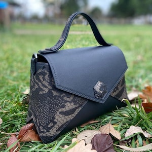 Puede incluir: Bolso de mano negro con cierre de solapa y asa superior. El bolso presenta un estampado de piel de serpiente en los laterales y el asa, con un cuerpo negro liso y un emblema decorativo en la solapa. El bolso está sobre hierba.
