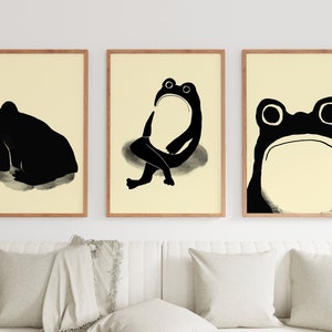 Three Matsumoto Hoji Frog Prints displayed in a triptych arrangement in a bright living space, each featuring a unique frog pose in bold black ink on cream paper, adding a touch of Japanese elegance