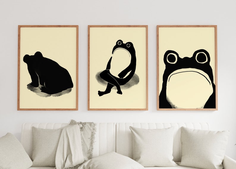 Three Matsumoto Hoji Frog Prints displayed in a triptych arrangement in a bright living space, each featuring a unique frog pose in bold black ink on cream paper, adding a touch of Japanese elegance