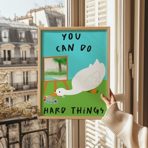 May include: A framed print with a white goose painting on a green background. The goose is holding a paintbrush and is near a small easel with a painting on it. The text on the print reads "You can do hard things."