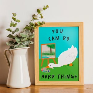 May include: A framed motivational poster with a blue background and a white goose painting a landscape. The text on the poster reads "You can do hard things."