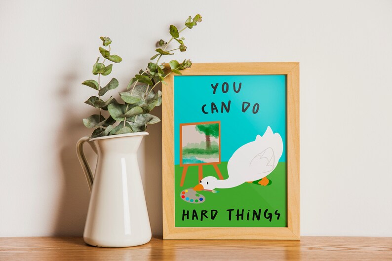 May include: A framed motivational poster with a blue background and a white goose painting a landscape. The text on the poster reads "You can do hard things."