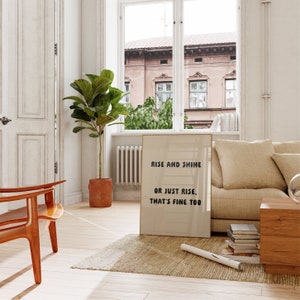 May include: A framed print with the text "Rise and Shine or Just Rise, That's Fine Too" leans against a beige sofa. The room features a wooden chair, a potted plant, and a window overlooking a building.