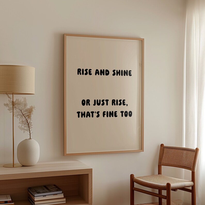 May include: A framed print with the text "RISE AND SHINE OR JUST RISE, THAT'S FINE TOO" in black, set against a cream background. The frame is a light wood color. The print is displayed in a room with neutral tones.