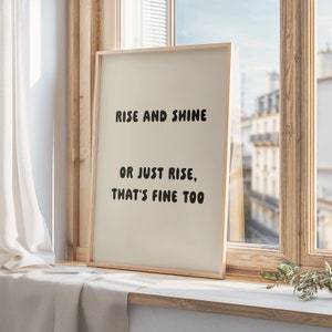 May include: A framed print with the text "RISE AND SHINE OR JUST RISE, THAT'S FINE TOO" in black, displayed on a windowsill. The frame is light-colored, and the background is a neutral beige. The window reveals a cityscape.