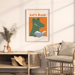 A framed "Let's Read" poster featuring a green alligator reading a book against an orange background, hanging in a cozy, sunlit reading corner with a wicker chair and a wooden console table.